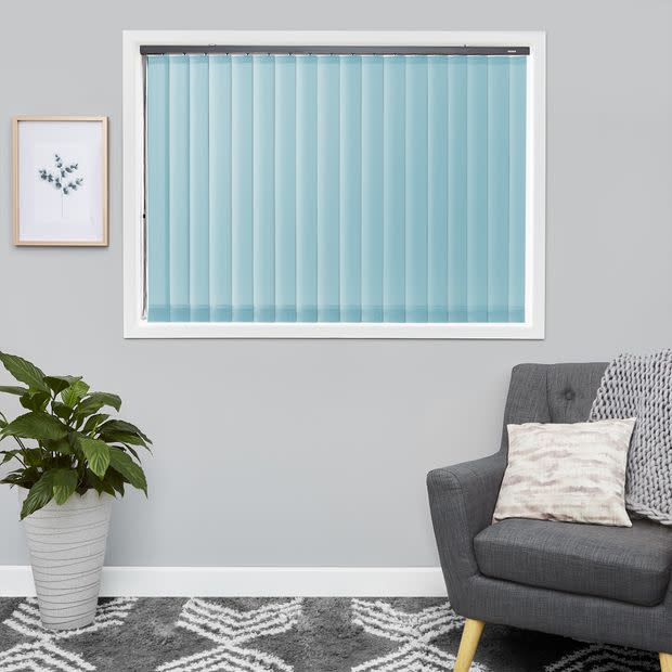 Light-blue vertical blinds fully closed across a rectangular window, filtering soft daylight, in a gray living room with a potted plant, framed botanical print, gray armchair, cushion and patterned rug.