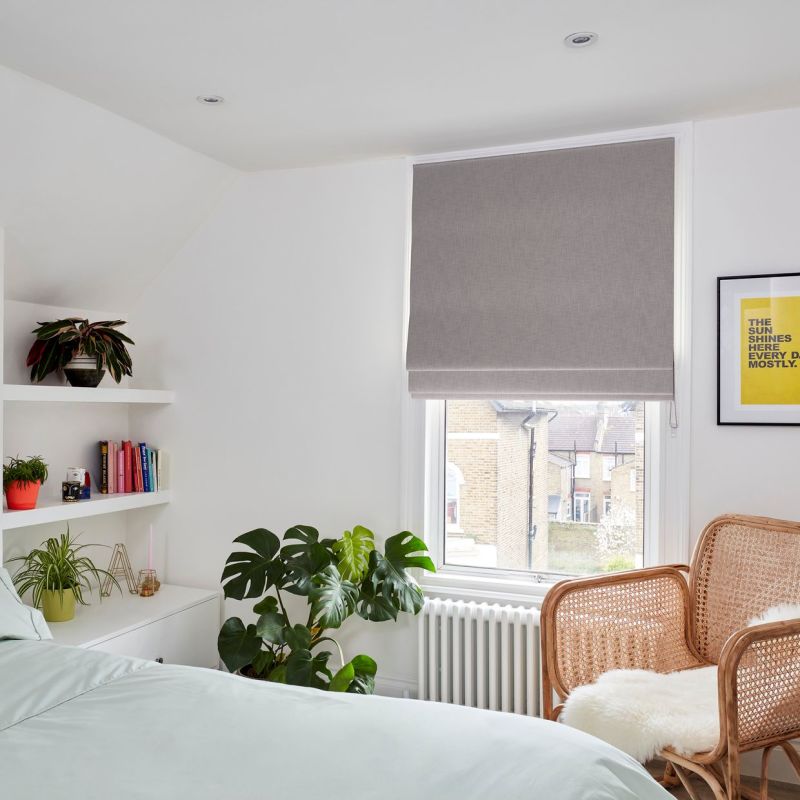 Grey fabric roman blind lowered over a sash window, filtering daylight; white bedroom with plants, wicker chair, radiator and a framed yellow print.

Transcription: THE SUN SHINES HERE EVERY DAY. MOSTLY.