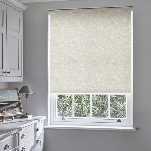 Beige patterned roller blind fully lowered, woven-arc motif filtering daylight; fitted inside a white-trim sash window in a softly lit gray study/dressing area, with cabinets, a lamp and painting beside.