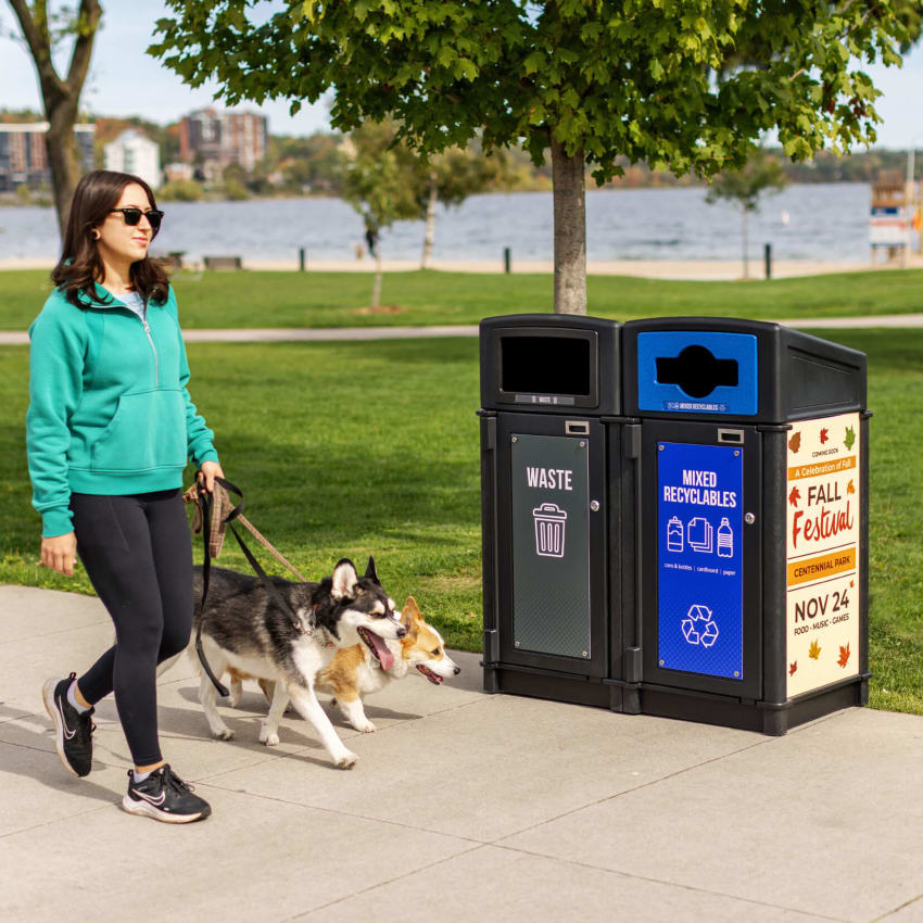 woman with cute dogs walking outdoor beside a double stream waste and recyclables bin with sloped lids and large body signs displaying collection information and a local event ad