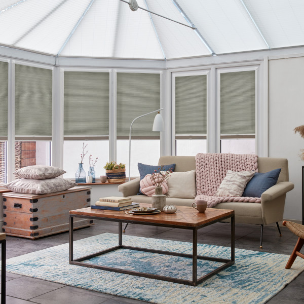 Pleated honeycomb blinds pulled down across tall conservatory windows, filtering soft daylight; the sunroom houses a beige sofa with chunky pink throw, wooden coffee table and a blue patterned rug.