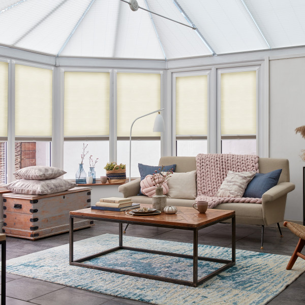 Cream honeycomb pleated blinds cover tall conservatory windows, lowered mid-height and softly diffusing daylight; cozy sunroom with beige sofa, pink knit throw, wooden coffee table and blue rug.