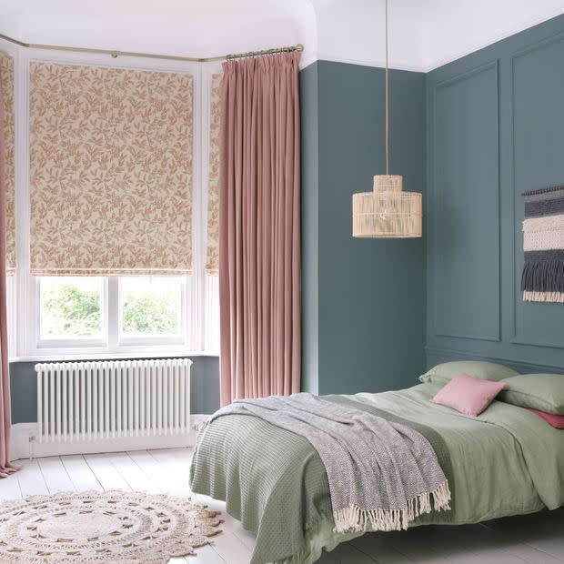 Floral-patterned roman blinds lowered over a bay window with long dusty-pink floor-length curtains drawn aside; they filter soft daylight into a teal-paneled bedroom with a green bed, woven pendant and round jute rug.