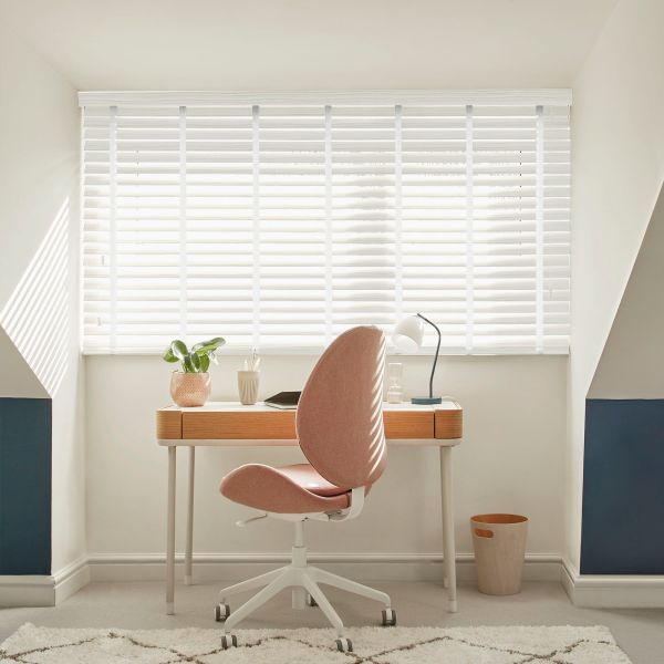White horizontal blinds covering a wide window, slats tilted to filter bright daylight; small attic-style home office with wooden desk, pink swivel chair, desk lamp, potted plant, neutral decor.