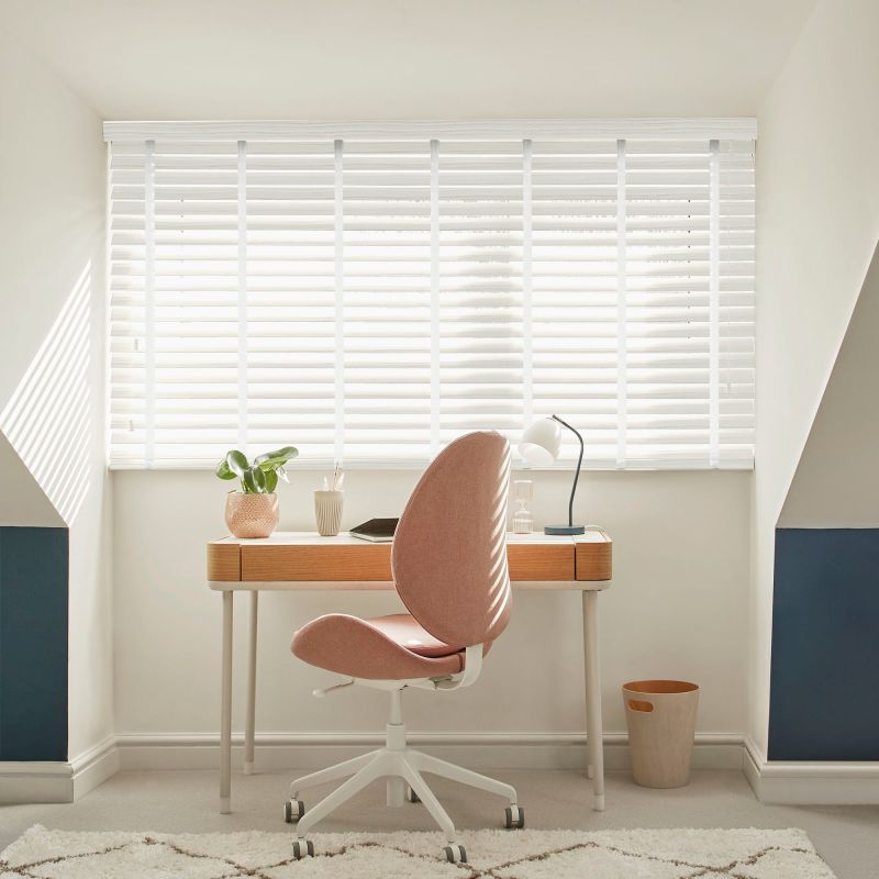 White horizontal blinds covering a wide window, slats tilted to filter bright daylight; small attic-style home office with wooden desk, pink swivel chair, desk lamp, potted plant, neutral decor.