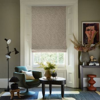 Textured beige Roman shade, lowered over a tall sash window with a narrow light gap at the bottom; filtering daylight into a cozy living room with armchair, carved coffee table, console and artwork.