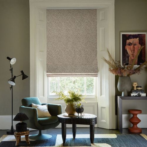 Textured beige Roman shade, lowered over a tall sash window with a narrow light gap at the bottom; filtering daylight into a cozy living room with armchair, carved coffee table, console and artwork.