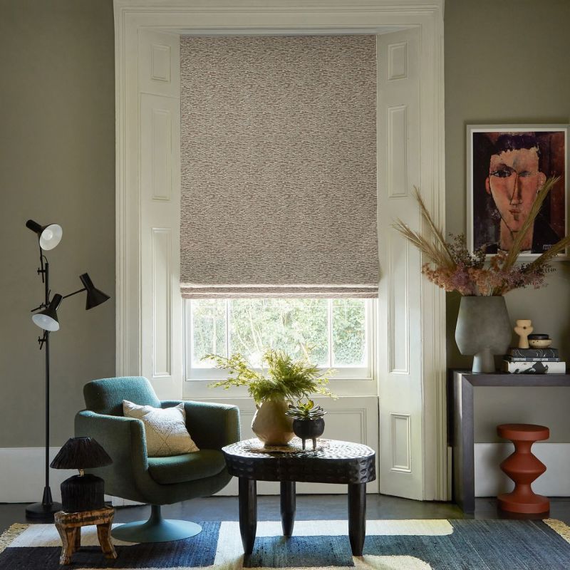 Textured beige Roman shade, lowered over a tall sash window with a narrow light gap at the bottom; filtering daylight into a cozy living room with armchair, carved coffee table, console and artwork.