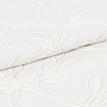 White embroidered fabric folded and overlapping, showing raised floral vine stitching and textured weave; close-up studio view with soft, even lighting emphasizing subtle shadows and fabric detail.