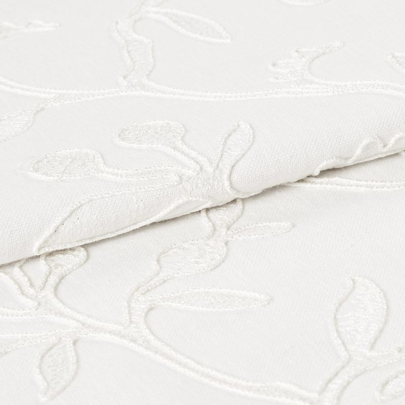 White embroidered fabric folded and overlapping, showing raised floral vine stitching and textured weave; close-up studio view with soft, even lighting emphasizing subtle shadows and fabric detail.
