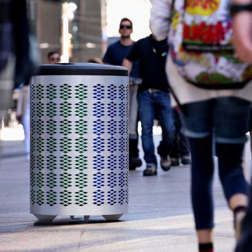 silver powder coated waste and recycling container with a black plastic lid on a busy city sidewalk with people walking by