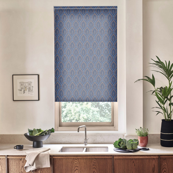 Blue patterned roller blind covers window above kitchen sink, blocking light; surrounding marble countertop holds faucet, bowl of leafy greens, potted plants and a framed sketch on pale wall.