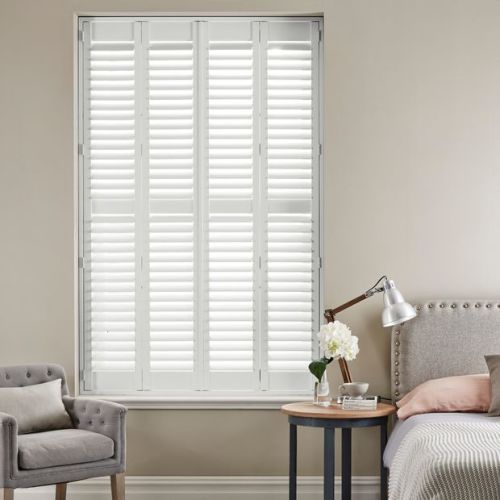 White plantation shuttersâ€”four hinged panels with horizontal louversâ€”cover a tall window; closed, filtering daylight into a neutral bedroom with upholstered bed, bedside table, lamp, armchair, and flowers.