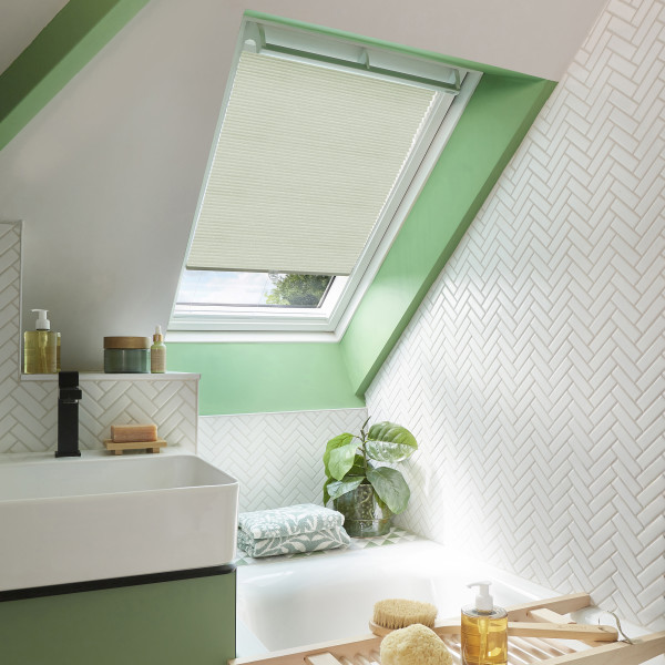 A cream pleated skylight blind, mostly lowered and filtering daylight through a slanted roof window; attic bathroom with bright green reveal, white herringbone tiles, sink, tub, plant and bath tray.