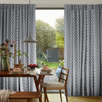 Gray patterned, floor-to-ceiling curtains, parted centrally and filtering soft daylight; a wooden dining table set with pancakes and fruit, pendant lamp above, chair and sunlit garden visible outside.