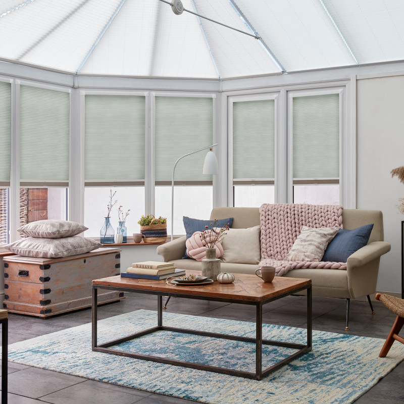 Pale cellular pleated blinds, lowered to mid-height across tall conservatory windows, filtering soft daylight; beige sofa with chunky pink throw and cushions; wooden coffee table on blue rug.