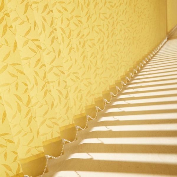 Vertical fabric blinds arranged along a side wall, scalloped bottoms linked by a bead chain, filtering strong sunlight into parallel stripes across a floor beside yellow leaf‑pattern wallpaper.