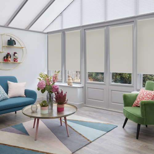 Cream roller blinds cover tall windows and triangular roof glazing, lowered to block upper light while leaving lower panes visible; sunlit living room with colorful sofas, round coffee table, rug.