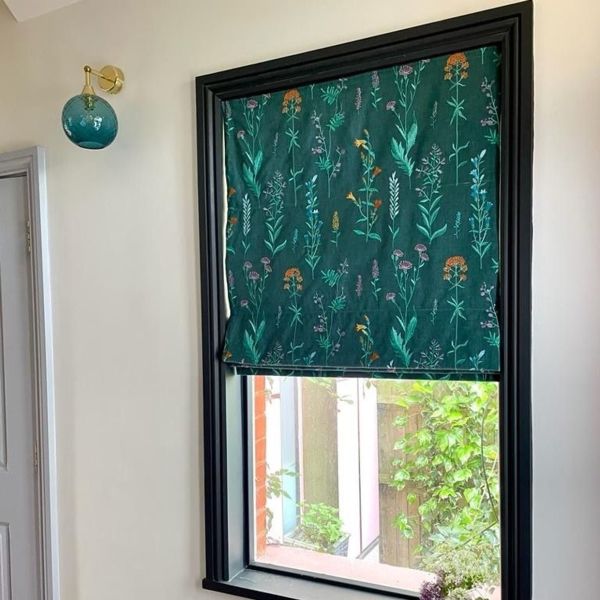 A floral-patterned teal Roman shade, lowered to cover the top half of a black-framed window and filtering light; nearby brass-and-glass wall sconce, pale hallway, and a small garden visible through the lower pane.
