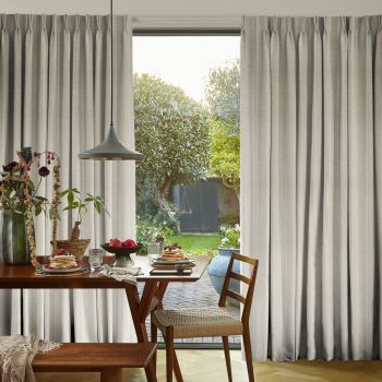Pleated beige floor-to-ceiling curtains, parted centrally to frame a glass door, filtering soft sunlight; dining table set with pancakes and fruit, pendant lamp overhead, view of a lush backyard.