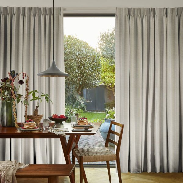 Pleated beige floor-to-ceiling curtains, parted centrally to frame a glass door, filtering soft sunlight; dining table set with pancakes and fruit, pendant lamp overhead, view of a lush backyard.