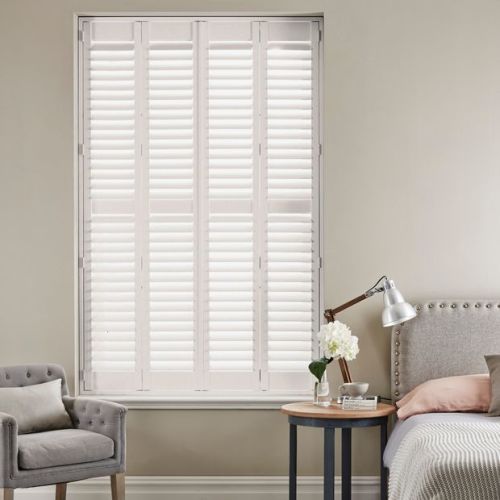 White plantation shutters covering a tall central window, louvered panels closed and filtering soft daylight; bedroom setting with upholstered bed, bedside table and lamp, plus an armchair against neutral walls.