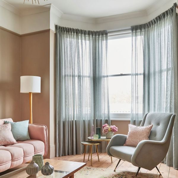 Sheer, floor-length pleated grey curtains hung across a bay window, gently filtering bright daylight; in a cozy living room with blush sofa, grey armchair, wooden floor, side tables and lamp.