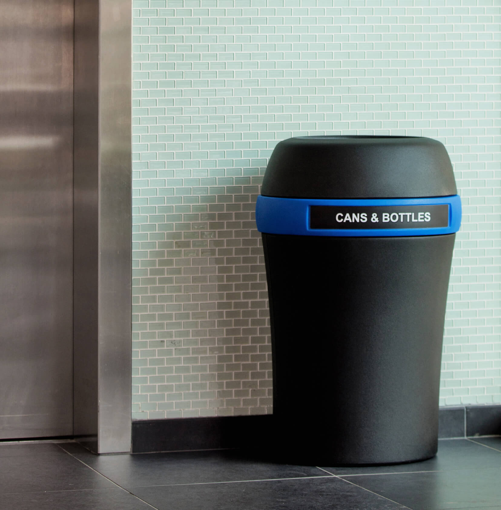 large black recycling bin with a blue band and cans & bottles label sitting in a hallway beside an elevator
