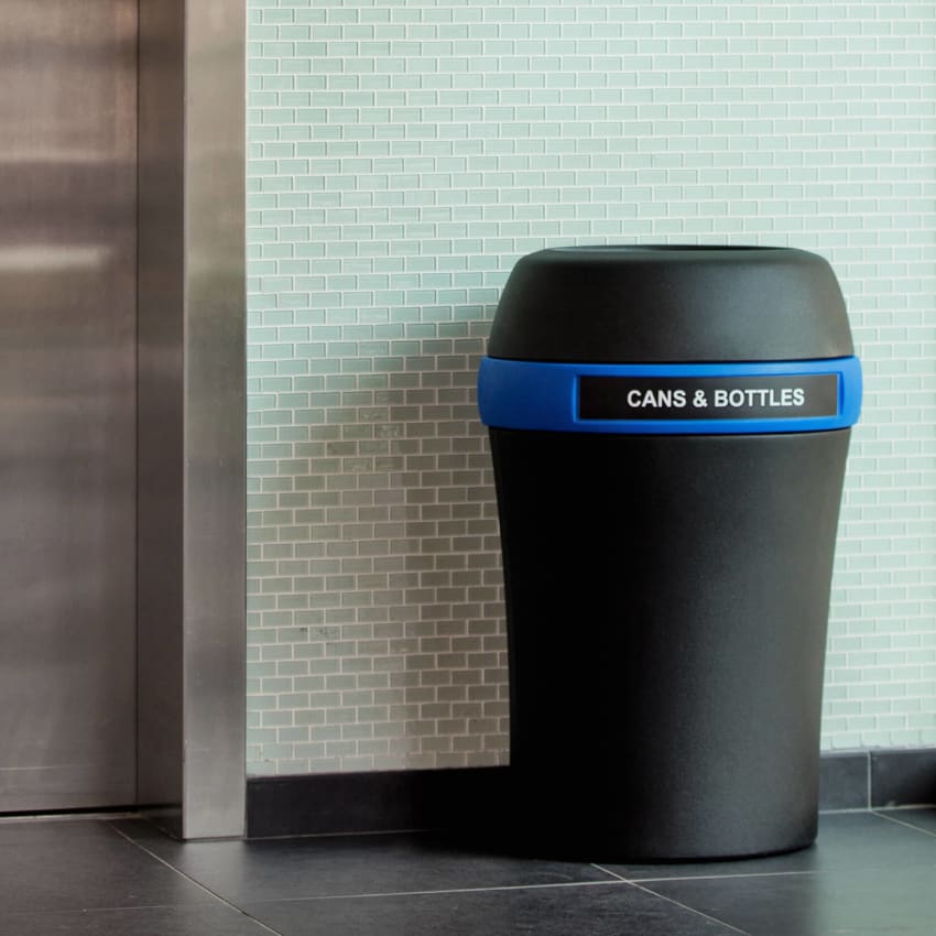 large black recycling bin with a blue band and cans & bottles label sitting in a hallway beside an elevator
