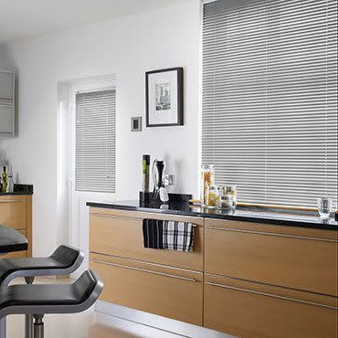 Silver horizontal Venetian blinds covering a large window and adjacent glazed door, thin metallic slats fully lowered; slats angled, filtering diffuse daylight; modern kitchen with light‑wood cabinets, black countertop, stools.