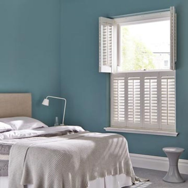 White plantation shutters on a sash window — upper panels folded open, lower panels closed with slats filtering light; small, softly lit bedroom with teal walls, bed and bedside lamp.