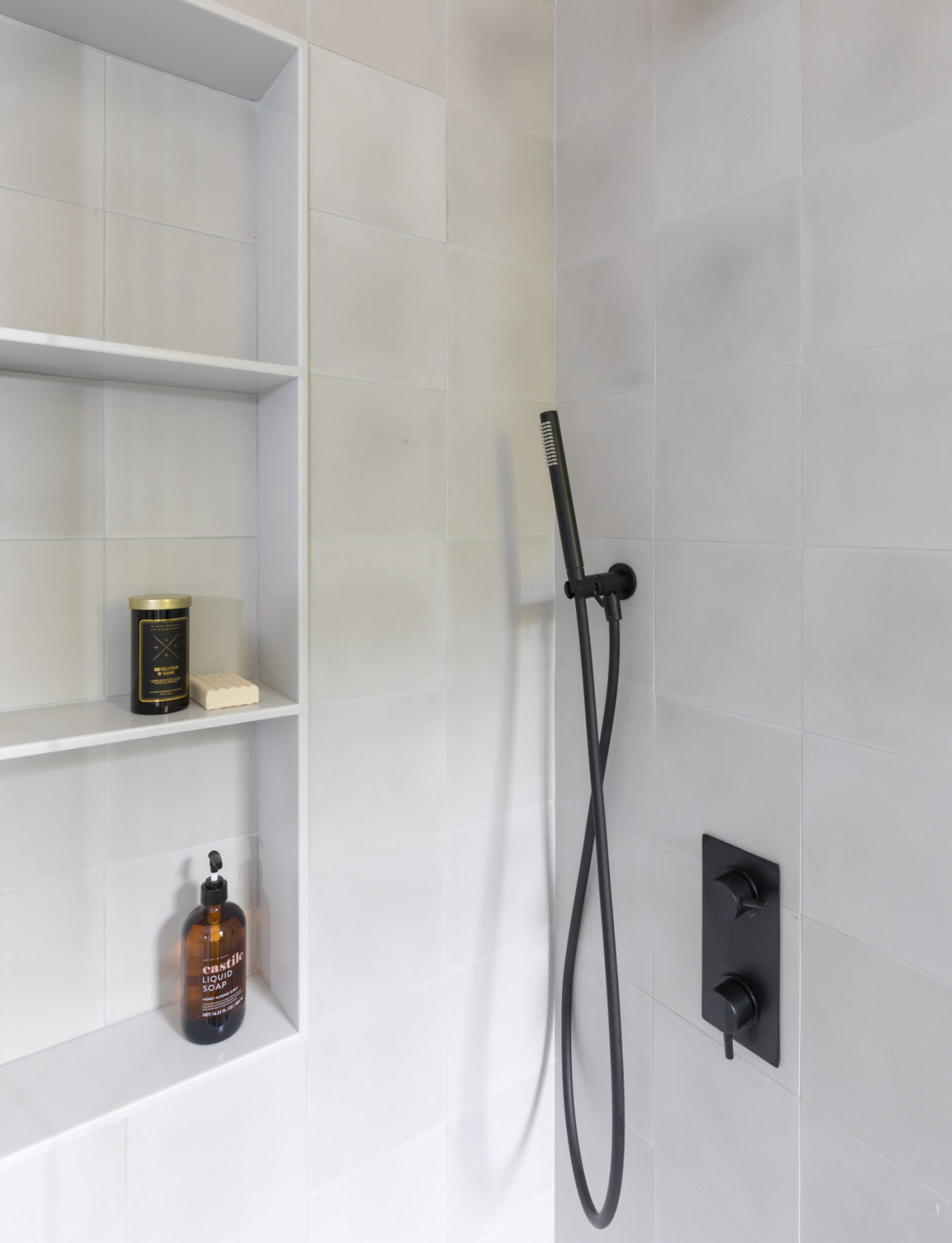 a white tiled shower with a black shower head and shelves.