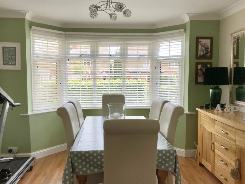 White horizontal Venetian blinds covering a bay window, partly closed and filtering daylight; a dining table with cream chairs faces them in a pale-green room with a wooden sideboard.

A & P