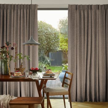 Floor-to-ceiling pleated grey curtains, parted centrally to frame a glass patio door, filtering soft morning light; dining table set with pancakes, fruit and pendant lamp, wooden chair, leafy backyard view.