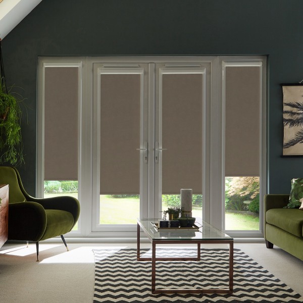 Floor-to-ceiling glass patio doors with beige roller blinds pulled down, framed by a dark wall; a glass coffee table on a black-and-white chevron rug sits between green armchairs, daylight outside.