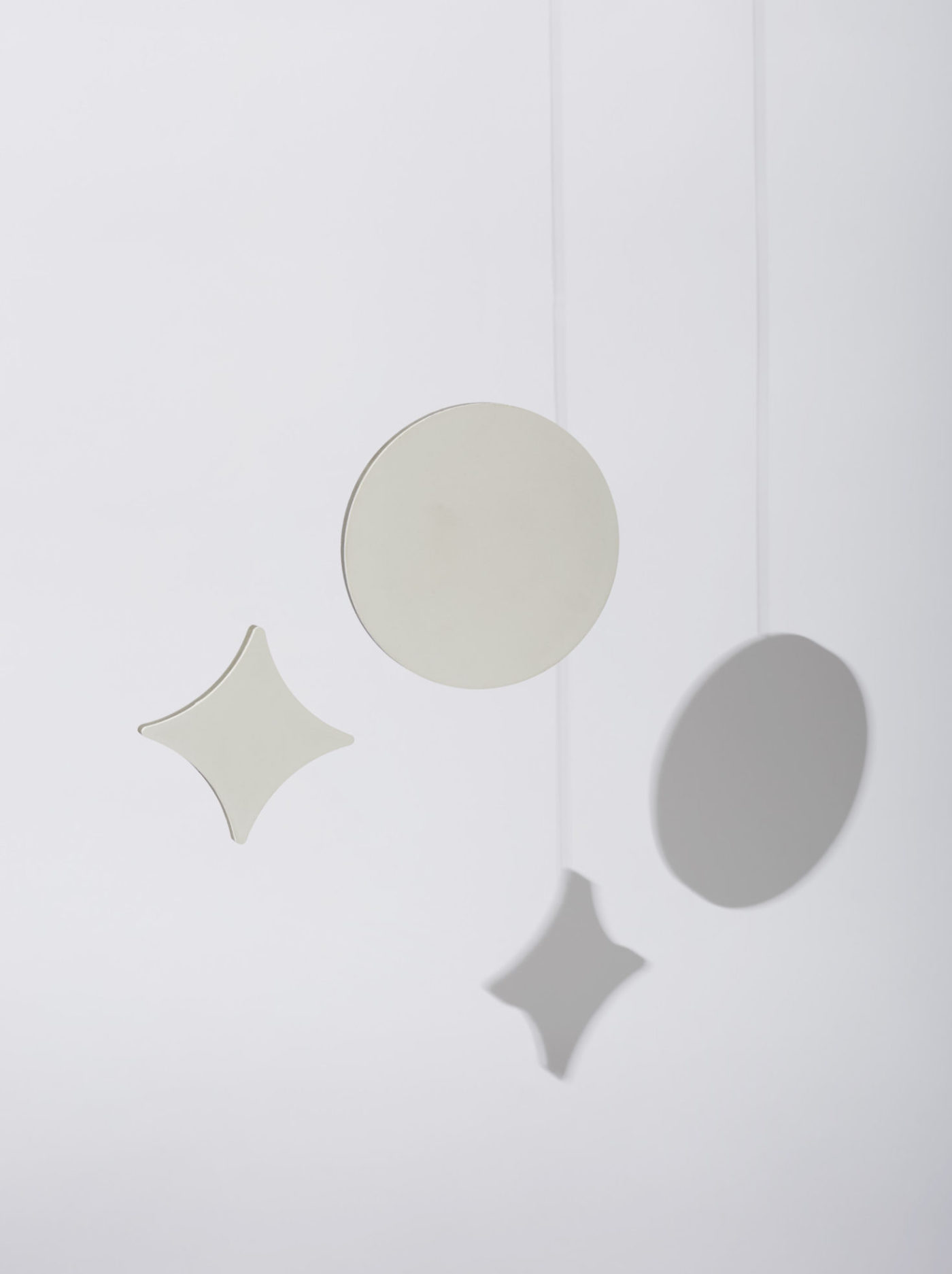 a white circle tile and white cross-shaped tile hanging from strings and casting shadows on a white wall.