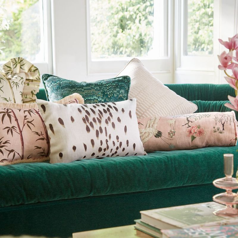 Emerald velvet sofa with assorted patterned throw pillows arranged along its backrest; bright bay windows flood the cozy living room with soft daylight and a leafy garden view.