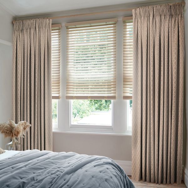 Beige patterned pinch-pleat curtains flank three lowered horizontal blinds whose angled slats filter soft daylight; small bedroom with a bed in the foreground and green foliage visible outside.