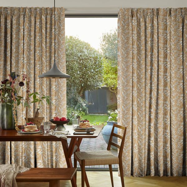 Printed, pleated, floor-length curtains with a beige-and-white leaf pattern, drawn slightly apart to frame a sliding glass door; allowing daylight into a cozy dining area with a set table and garden view.