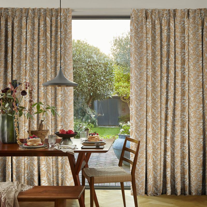 Printed, pleated, floor-length curtains with a beige-and-white leaf pattern, drawn slightly apart to frame a sliding glass door; allowing daylight into a cozy dining area with a set table and garden view.