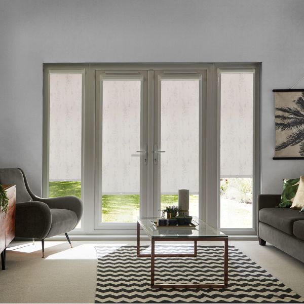 White patterned roller blinds on central French doors and sidelights, lowered to filter daylight and reveal a lawn strip beneath; living room with grey seating, coffee table and chevron rug.