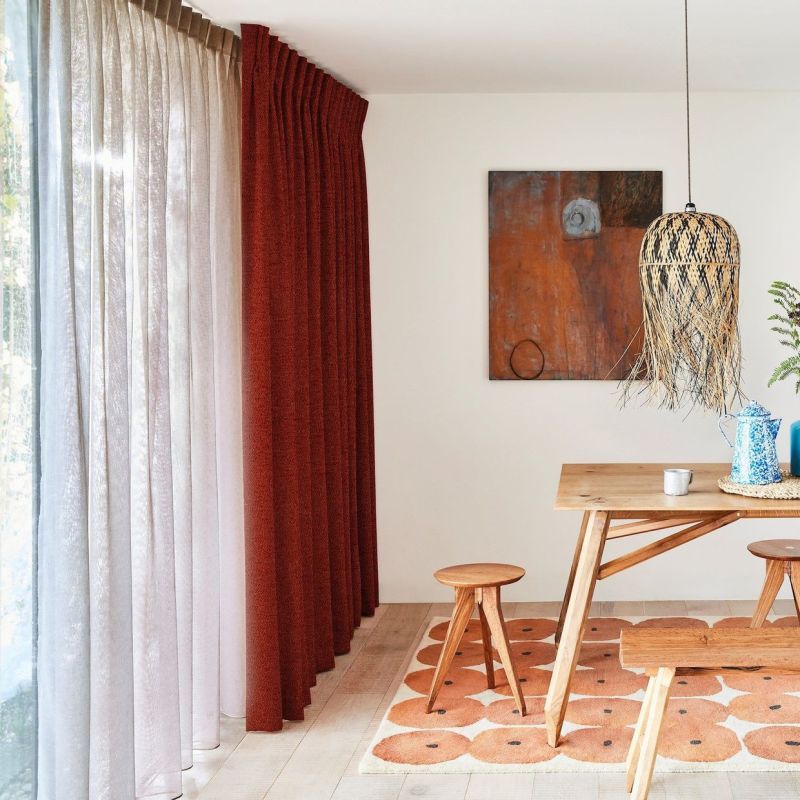 Floor-to-ceiling sheer white panels with pleated rust-red drapes at left, rust curtain drawn while sheer filters daylight; dining nook with wooden table, stools, woven pendant, abstract wall art, patterned rug.