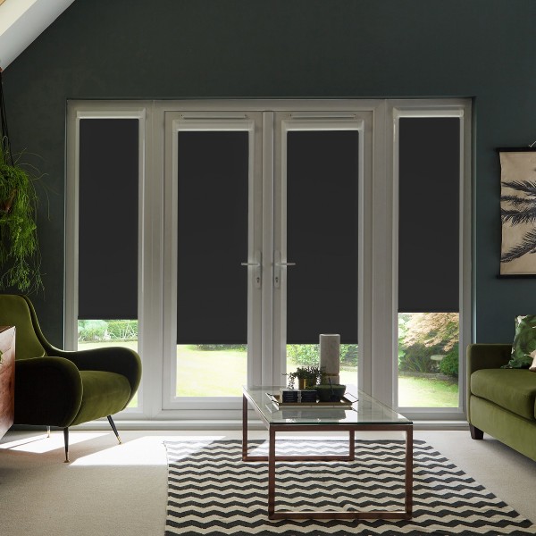 French glass doors and adjoining windows feature dark roller blinds pulled down to mid-height, framing a sunlit garden beyond; living room foreground with green chairs, glass coffee table and rug.