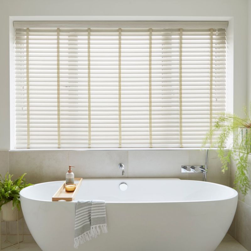 Cream horizontal slatted blinds with vertical ladder tapes cover a recessed window, closed and softly filtering daylight, above a freestanding white bathtub in a neutral, plantâ€‘accented bathroom.