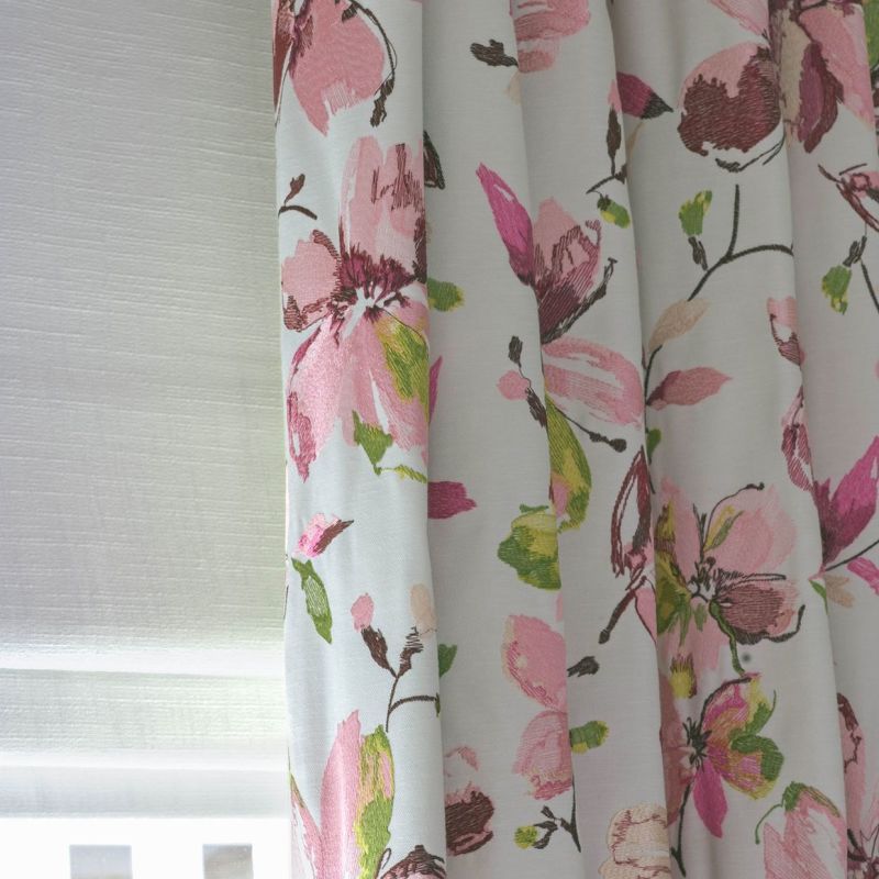 Floral fabric curtain â€” pleated and drawn at right with large pink and green blossoms â€” partially covering a lowered white roller blind, filtering soft daylight in a neutral-walled interior.
