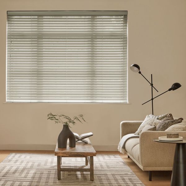 Horizontal white Venetian blinds cover a wide window, closed and softly filtering daylight; living room with beige sofa, wooden coffee table, sculptural vase and open book, patterned rug, floor lamp.