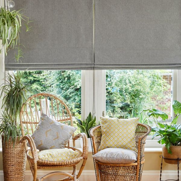 Grey fabric Roman blinds, lowered with soft folds, partially filtering bright daylight through a narrow lower gap; sunlit sitting area with wicker chairs, patterned cushions, potted plants and garden view.