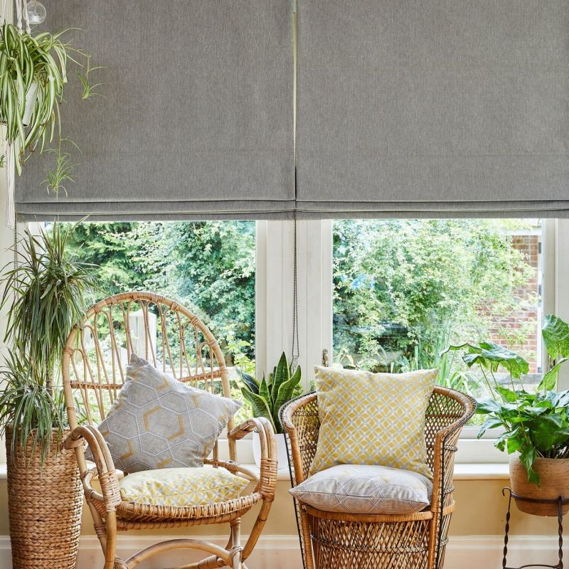 Grey fabric Roman blinds, lowered with soft folds, partially filtering bright daylight through a narrow lower gap; sunlit sitting area with wicker chairs, patterned cushions, potted plants and garden view.