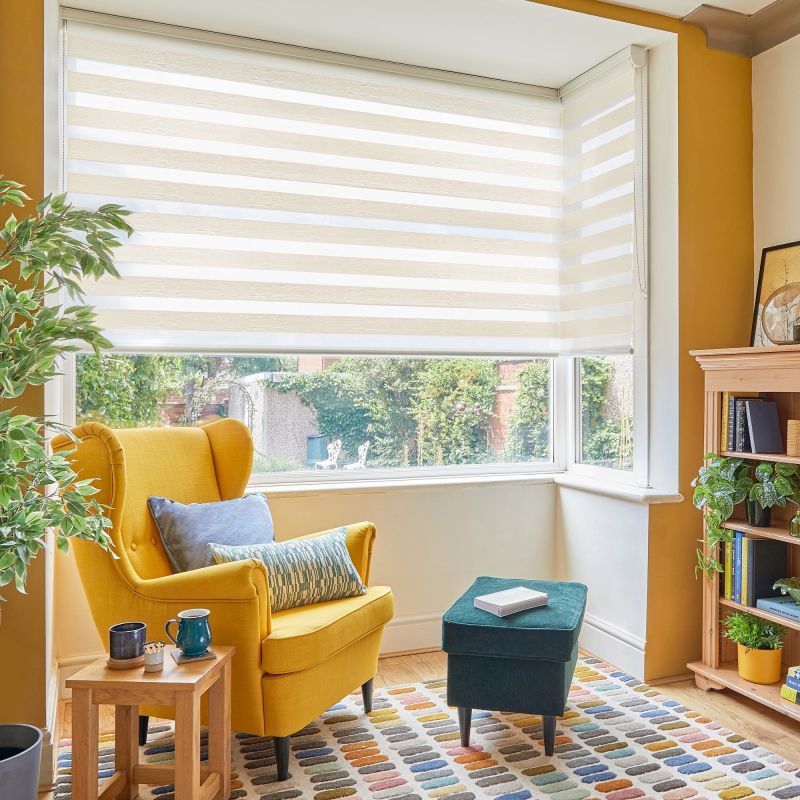 Zebra roller blind, lowered across a bay window with alternating opaque and sheer stripes, partially closed and filtering daylight; cozy living-room corner with mustard wingback chair, green ottoman, rug and plants.
