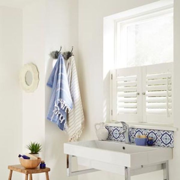 White café-style plantation shutters covering the lower window, slatted panels slightly ajar, filtering sunlight into a bright bathroom with a white wall-mounted sink, patterned tile backsplash, hanging towels.